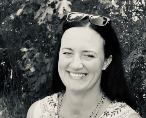 Ania Behr auf einem Schwazweißbild. Sie llacht und steht vor einem Strauch-Hintergrund.